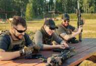 Positive young men in body armors sitting at wooden table and chatting while reloading guns